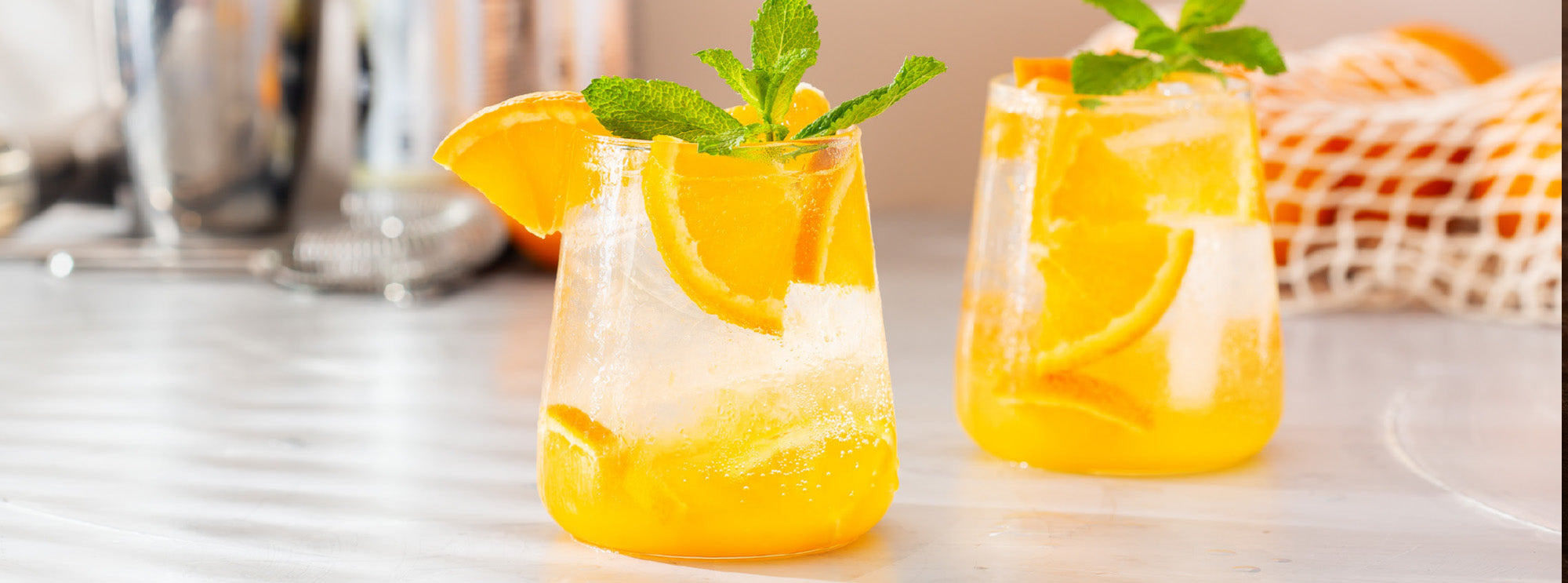 Two glasses of orange juice with lemon slices and mint leaves on a white surface.